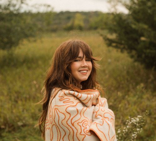 Artist Madison Holler of Rubinski Works outdoors in a field, wrapped in an orange patterned blanket