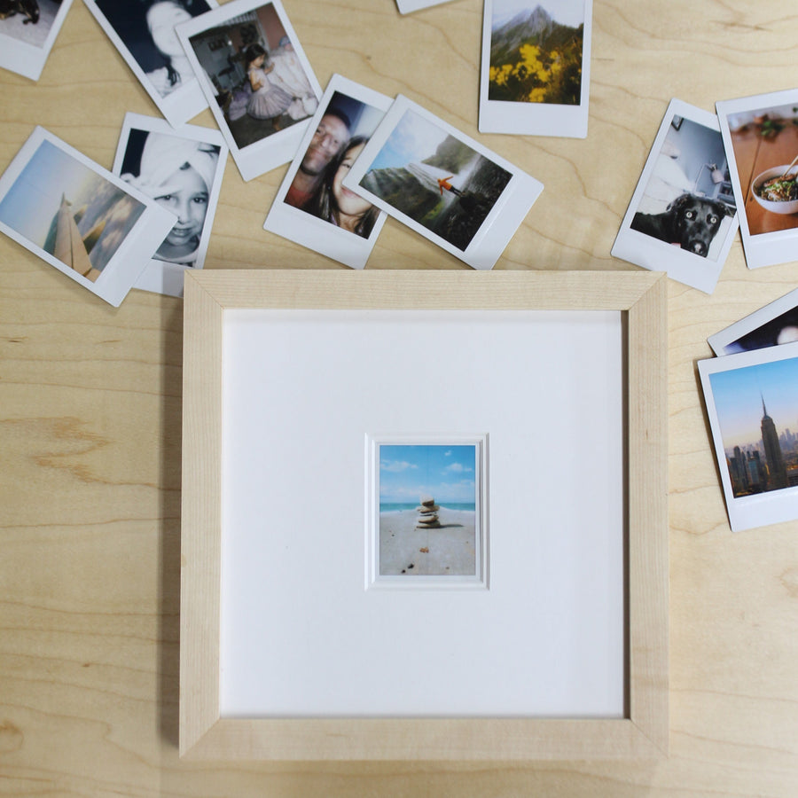 Maple frame holding a single Instax Mini photo, surrounded by loose instant photos on a wood table