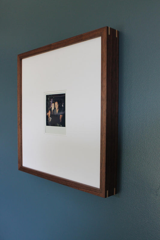 Walnut gallery frame angled on a teal wall with a wide white mat and a Polaroid photo