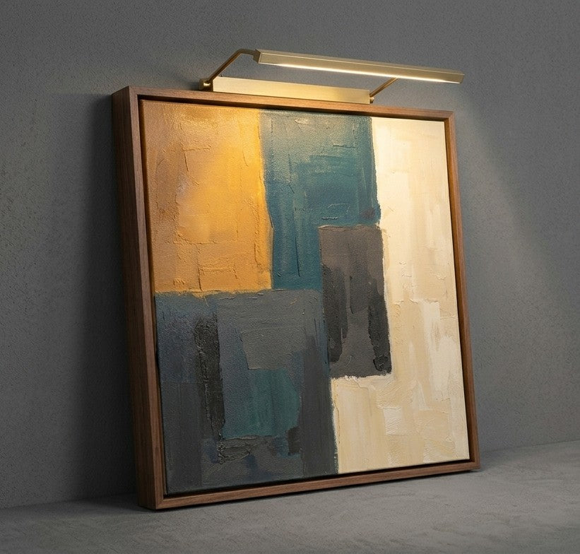 A framed abstract canvas painting with geometric blocks of mustard yellow, teal, gray, and cream, displayed in a warm wood frame against a textured gray wall with a modern wall light above it.
