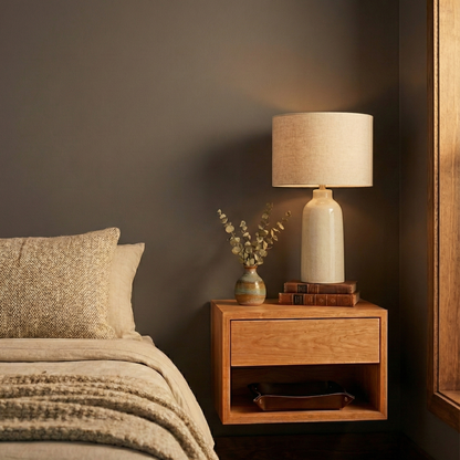 Wooden nightstand with lamp and decor items next to a bed with pillows and blanket.